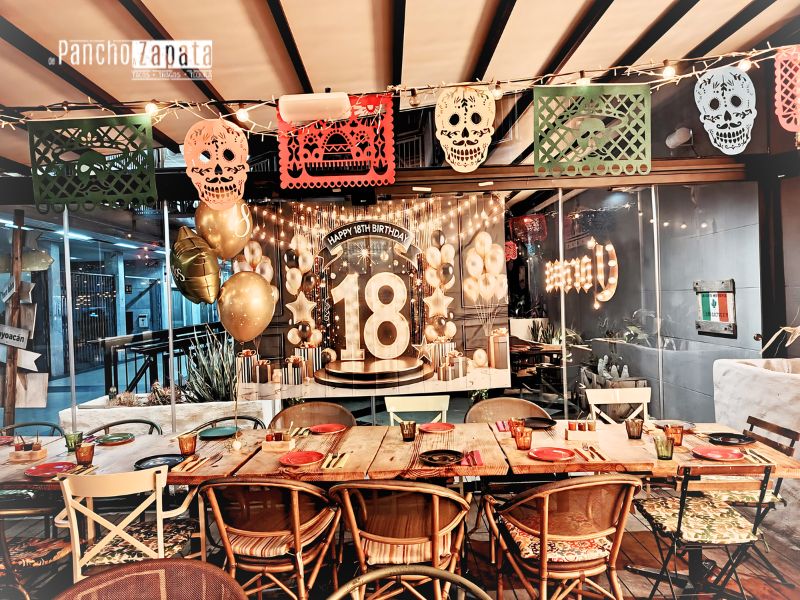 Mesa larga decorada para una fiesta de 18 cumpleaños en De Pancho a Zapata, con papel picado y globos dorados